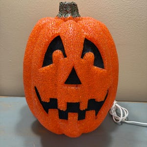 May include: A bright orange jack-o'-lantern pumpkin decoration with a textured, glittery surface. It features a carved face with triangular eyes, a triangular nose, and a jagged mouth with black cutouts. A green stem sits on top and a white cord.