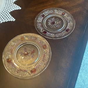May include: Two clear glass plates with a raised design of fruit and leaves. The plates have a pink tint and are on a dark brown wooden surface.
