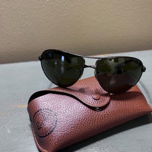 May include: Dark-framed sunglasses with green lenses rest on a brown leather case. The case has a snap closure and a debossed logo. The sunglasses have a classic aviator style, with a double bridge and a sleek, modern design.