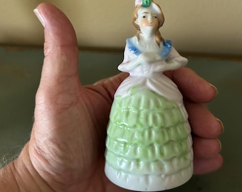 Peut inclure: Petite figurine vintage en porcelaine représentant une femme dans une longue robe verte à volants et un corsage blanc. La figurine porte un chapeau blanc avec un accent floral rose et vert et une bordure bleue.