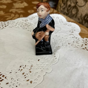 May include: A ceramic figurine of a boy wearing a blue scarf and holding a brown stick. The figurine is sitting on a black base.