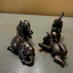May include: Four bronze-colored elephant figurines in various poses. Two are lying down, one is standing, and another is balancing on top of two others. The figurines have detailed textures and are arranged on a green surface.