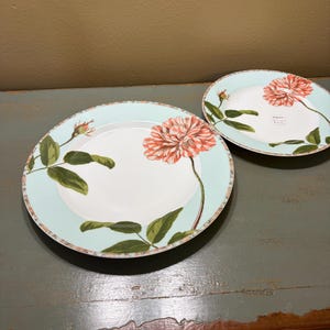Puede incluir: Dos platos de cerámica blanca con borde azul claro y diseño floral. Los platos presentan una gran flor de color coral con hojas y capullos verdes. Los platos están sobre una superficie de madera envejecida.