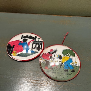 May include: Two vintage round metal buttons with red rims. One depicts a person kneeling by a bed with the text "ASK OF GOD". The other shows figures working in a garden with the text "THAT SHALL YE ALSO REAP".
