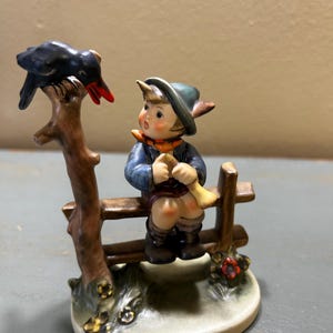 Peut inclure: Figurine en céramique représentant un garçon jouant d'un cor sur une clôture en bois. Un oiseau noir au bec rouge est perché sur une branche d'arbre. Le garçon porte une veste bleue, un chapeau et une écharpe orange. La base est ovale.