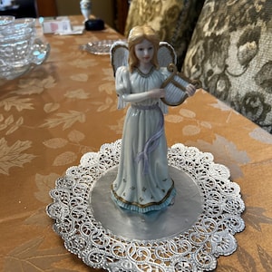 May include: A porcelain angel figurine with blonde hair and wings, holding a harp. The figurine is standing on a silver doily.