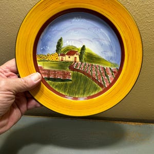 Puede incluir: Un plato de cerámica decorativo con una escena de paisaje pintada a mano. El plato tiene un borde amarillo con un borde interior marrón. La imagen central representa una escena rural con una casa, árboles y un campo, bajo un cielo azul.