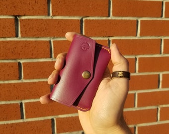 May include: A burgundy leather key holder with a yellow stitch detail and a brass button closure. The key holder is held in a hand with a brown leather bracelet.