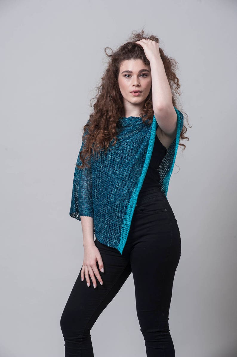 Puede incluir: Una mujer con el pelo largo y rizado casta&ntilde;o lleva un poncho de punto azul turquesa con un top negro de tirantes y unos vaqueros negros.