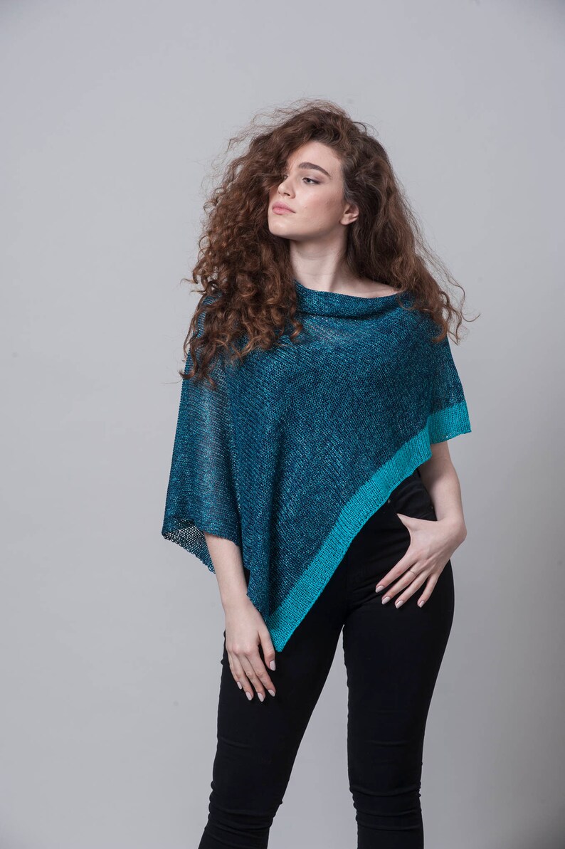 Puede incluir: Una mujer con un poncho de punto azul turquesa y turquesa de corte holgado y dobladillo ligeramente asim&eacute;trico. Lleva pantalones vaqueros negros y tiene el pelo largo y rizado casta&ntilde;o.
