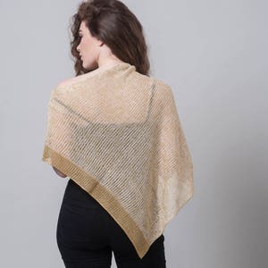 Puede incluir: Una mujer con un poncho de punto beige y dorado, un top negro y unos vaqueros negros.