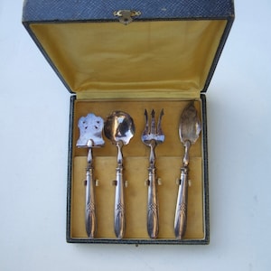 Peut inclure: Un ensemble de service argenté vintage, comprenant une pelle à gâteau, une cuillère, une fourchette et une spatule, présenté dans un coffret doublé bleu et or. L'ensemble présente des manches ornés et date probablement du début du XXe siècle.