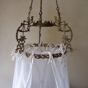 May include: An antique brass hanging canopy frame with decorative floral and bird-like accents. The frame supports a white, sheer fabric curtain, tied with white fabric. The canopy is suspended from chains.