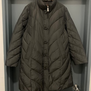 May include: A dark gray, quilted winter coat with a high collar and button closure. The coat is hanging on a wooden hanger and features a chevron pattern. The coat has two front pockets and a zipper closure.