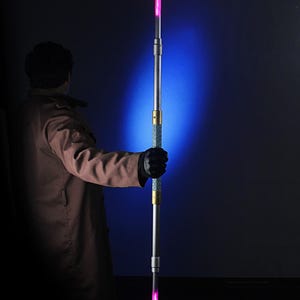 May include: A double-bladed lightsaber with a silver handle and pink glowing blades. The handle has gold and blue accents. The lightsaber is held against a dark background with a blue spotlight.