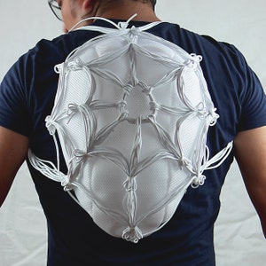 May include: A white, web-like structure made of knotted cords is worn on the back of a person. The structure is shaped like a dome and has a circular opening in the center.