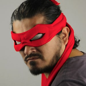 May include: A vibrant red eye mask and head wrap, similar to a superhero or comic book character. The mask features mesh eye coverings and is made of a soft material. The person is wearing a grey top. The overall look is striking.