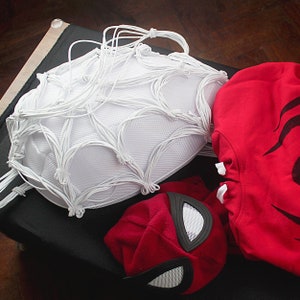 May include: A white web-like mask with a red and black mask underneath. The masks are on a black surface.