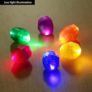 May include: Six colorful, faceted, gem-shaped, light-up toys. The gems are red, green, purple, yellow, blue, and orange. The gems are arranged in a circle on a brown surface.