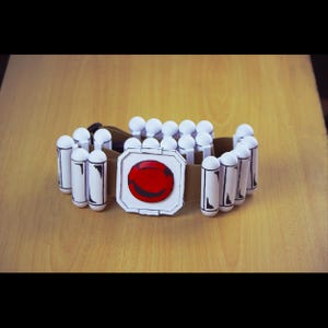 Puede incluir: Un cinturón blanco con un círculo rojo y una cara sonriente negra en la hebilla. El cinturón está hecho de espuma y tiene una correa marrón.