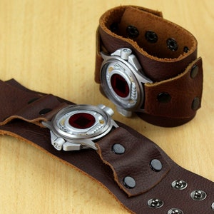 Può includere: Due orologi da polso in pelle marrone con quadranti color argento e centri rossi. Gli orologi hanno un'estetica steampunk, con ingranaggi e rivetti visibili. Un orologio è aperto, l'altro è chiuso.