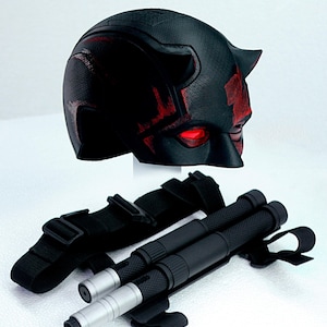 Peut inclure: Casque de super-héros noir et rouge avec des cornes et des lentilles oculaires rouges incandescentes. En dessous, deux bâtons noirs avec des embouts argentés, fixés par des sangles et des boucles noires. Sur fond blanc.