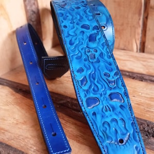 Pode incluir: Uma correia de guitarra de couro azul com um design de caveira e chamas. A correia é feita de couro de alta qualidade e apresenta um padrão em relevo detalhado. Uma extremidade de correia azul correspondente também é visível.