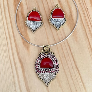 May include: A silver choker necklace with a red stone pendant. The pendant is surrounded by smaller red stones and has a floral design. The earrings are also red stone with a floral design.