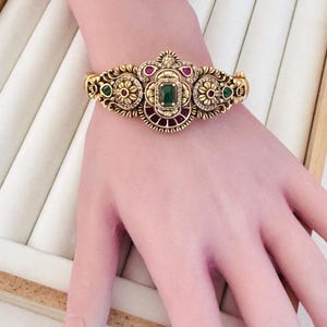 May include: A gold-toned metal cuff bracelet featuring an ornate, textured design with a central rectangular green stone, accented by smaller circular green and red stones, and embedded with tiny clear crystals along the decorative metalwork.