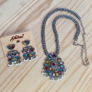 May include: A silver necklace with a peacock pendant featuring colorful stones and pearl accents. Matching earrings with similar design and stones are also included. The necklace has a chain with a leaf-like pattern.