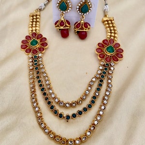 Puede incluir: Un collar de oro de varias capas con detalles de flores rojas y verdes y pendientes a juego. El collar tiene tres capas de cadena de oro con piedras transparentes y verdes.