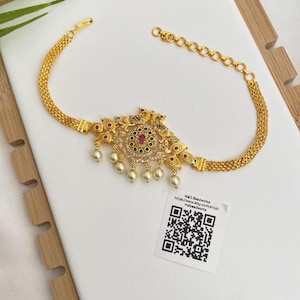 May include: Gold-toned choker necklace with a central floral design, adorned with red, green, and clear gemstones, and dangling pearls. The necklace features a chain-link design and a small tag with a QR code.
