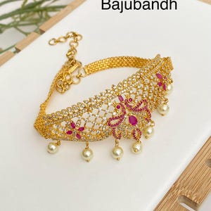 May include: Gold-toned armlet with the word "Bajubandh" in black. The armlet features a filigree design with clear and ruby-colored stones, and dangling faux pearls. The armlet is on a white surface.