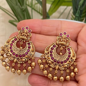 May include: A pair of gold earrings with a crescent moon design and pink stones. The earrings have a dangling design with small gold balls.