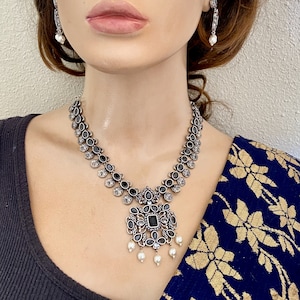 May include: A silver necklace with a black stone pendant and pearl accents. The necklace is made of metal and has a traditional design.