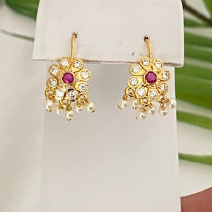 May include: Gold-toned floral earrings with a central red gemstone and surrounding clear stones. Small white pearls dangle from the bottom of each earring. The earrings are displayed on a white surface.