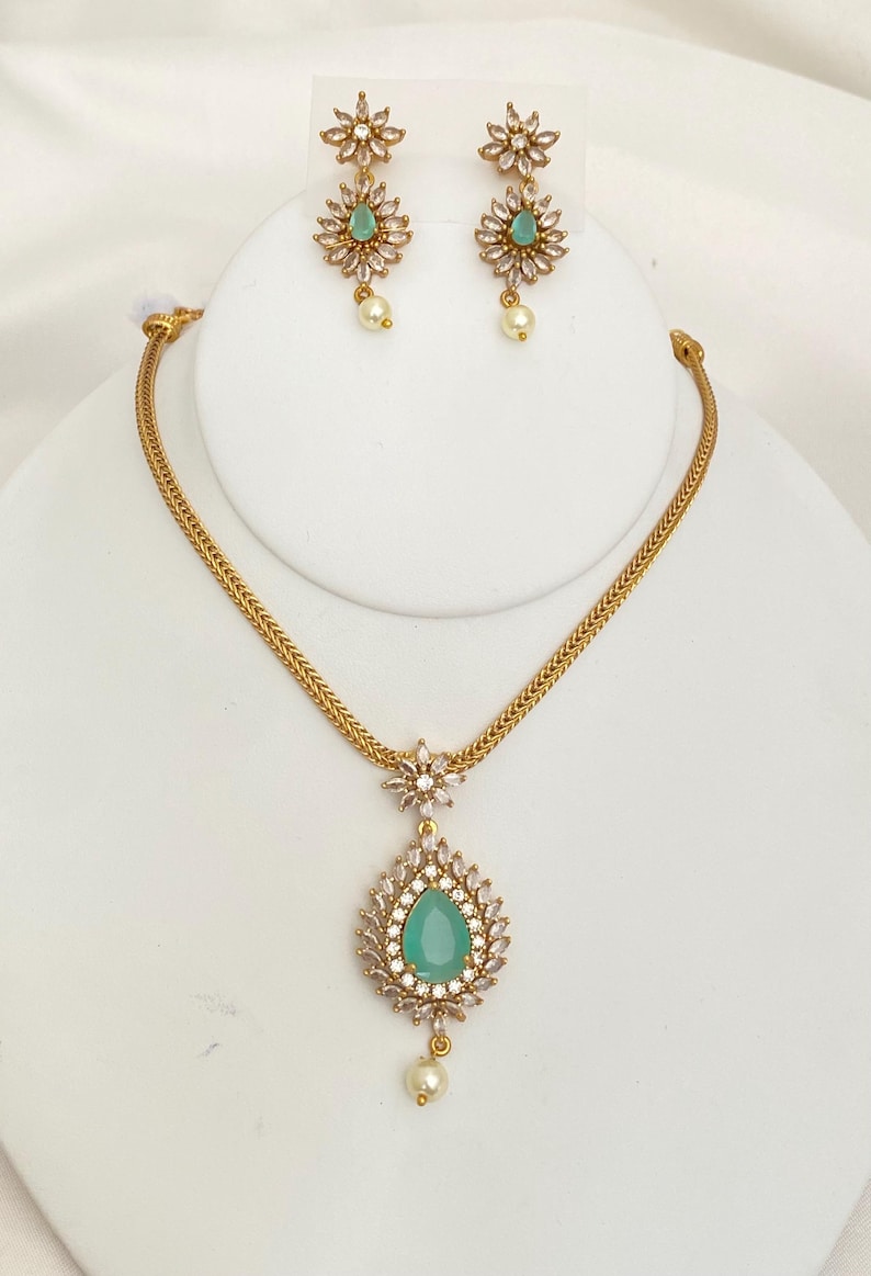 May include: A gold-toned jewelry set featuring a necklace and earrings. The necklace has a teardrop-shaped pendant with a teal center stone, surrounded by clear crystals, and a pearl drop. The earrings mirror the pendant design with a flower-shaped top and pearl drop.