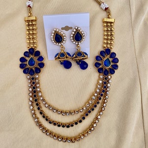 Puede incluir: Un collar de oro con tres hileras de piedras azules y blancas. El collar tiene dos colgantes de flores azules y pendientes a juego con piedras azules en forma de l&aacute;grima.