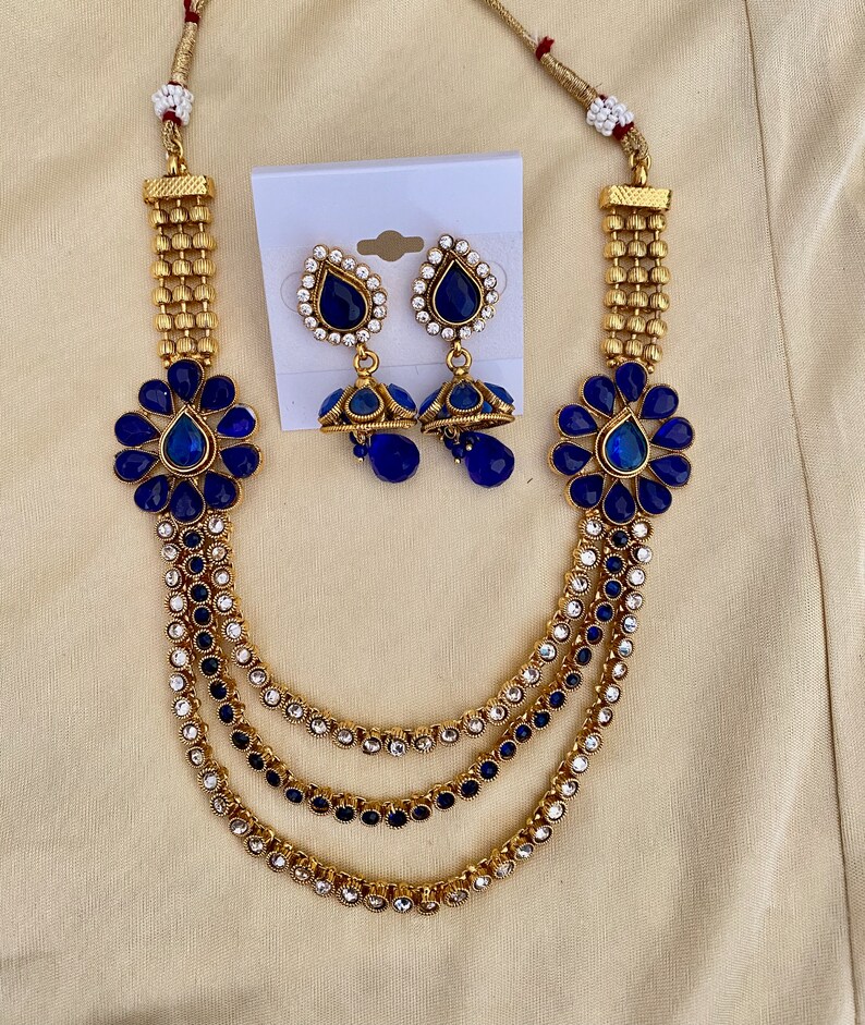 Puede incluir: Un collar de oro con tres hileras de piedras azules y blancas. El collar tiene dos colgantes de flores azules y pendientes a juego con piedras azules en forma de l&aacute;grima.