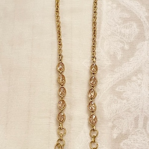 May include: Two gold-colored jewelry chains with oval-shaped, clear gemstones. Each chain has a hook at the top and a decorative loop at the bottom. The chains are displayed on a white, textured surface.