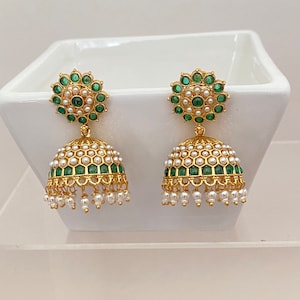 May include: A pair of gold-colored earrings with a floral design at the top, featuring green and white gemstones. The bell-shaped body is adorned with white gemstones and small dangling pearls. The earrings are displayed on a white surface.