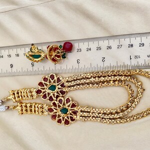 Puede incluir: Un collar de oro con tres cadenas y dos colgantes florales rojos y verdes. El collar se mide con una regla, mostrando que tiene aproximadamente 71 cm de largo.