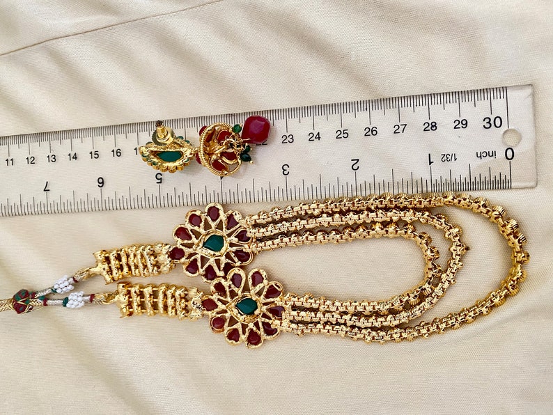 Puede incluir: Un collar de oro con tres cadenas y dos colgantes florales rojos y verdes. El collar se mide con una regla, mostrando que tiene aproximadamente 71 cm de largo.