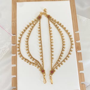 May include: A pair of gold-toned metal anklets featuring a multi-layered chain design with small square-shaped beads and two circular floral accents at each end, each centered with a small round red stone, connected by a central clasp mechanism.