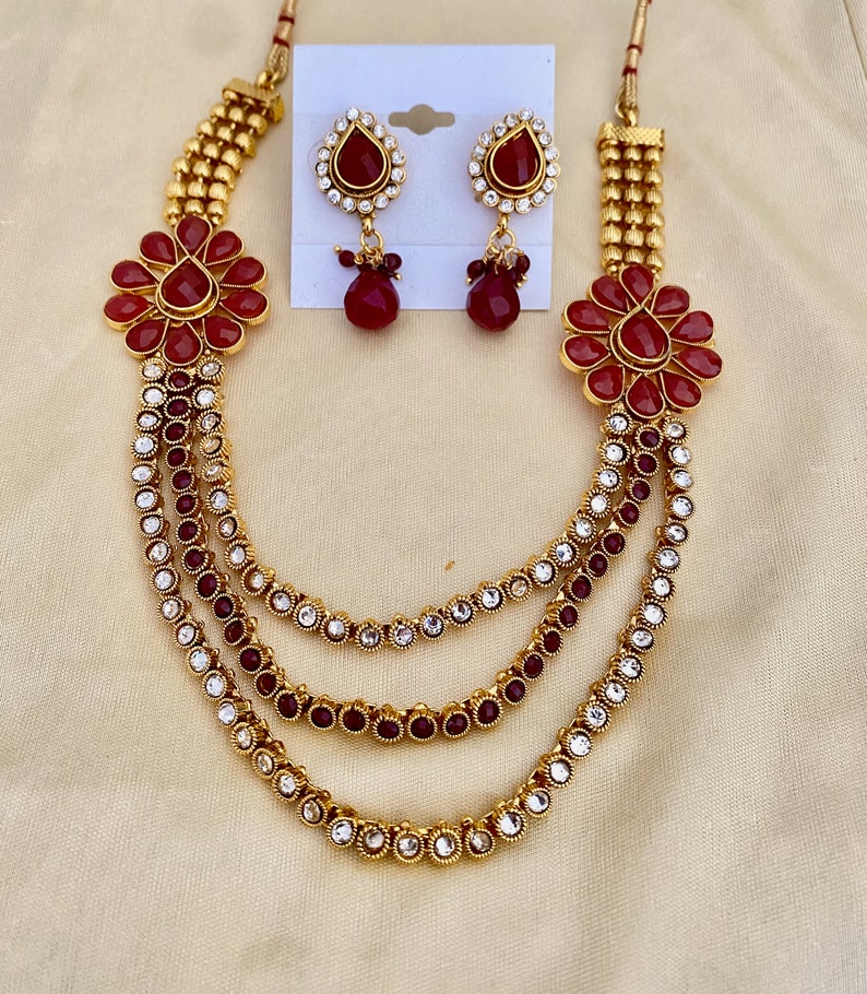 Puede incluir: Un collar de oro con cuatro hileras de piedras rojas y transparentes, dos colgantes de flores rojas a juego y un par de pendientes de piedras rojas y transparentes.
