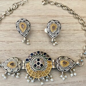 May include: A silver and gold necklace with a circular pendant featuring blue and pink stones. The pendant is surrounded by smaller teardrop-shaped pendants with gold accents. The necklace is paired with matching earrings.