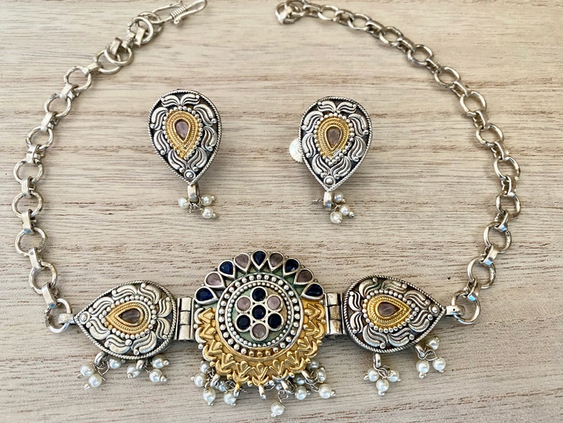 May include: A silver and gold necklace with a circular pendant featuring blue and pink stones. The pendant is surrounded by smaller teardrop-shaped pendants with gold accents. The necklace is paired with matching earrings.