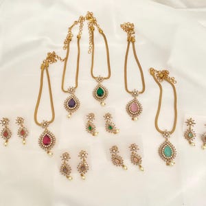 May include: A collection of gold-toned necklaces and matching earrings. Each necklace features a teardrop-shaped gemstone pendant in various colors, including red, purple, green, pink, and teal. The earrings have a floral design with a small pearl.