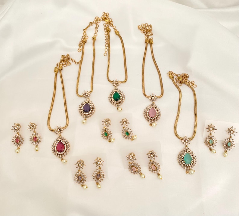 May include: A collection of gold-toned necklaces and matching earrings. Each necklace features a teardrop-shaped gemstone pendant in various colors, including red, purple, green, pink, and teal. The earrings have a floral design with a small pearl.