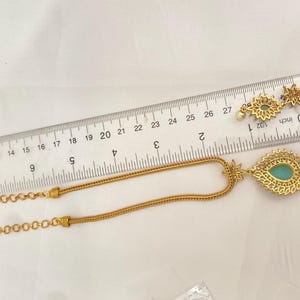 May include: Gold-toned necklace and earrings set. The necklace features a teardrop pendant with a teal-colored stone and a delicate chain. The earrings have a similar design, with a starburst pattern and a small pearl. A ruler is placed next to the jewelry.